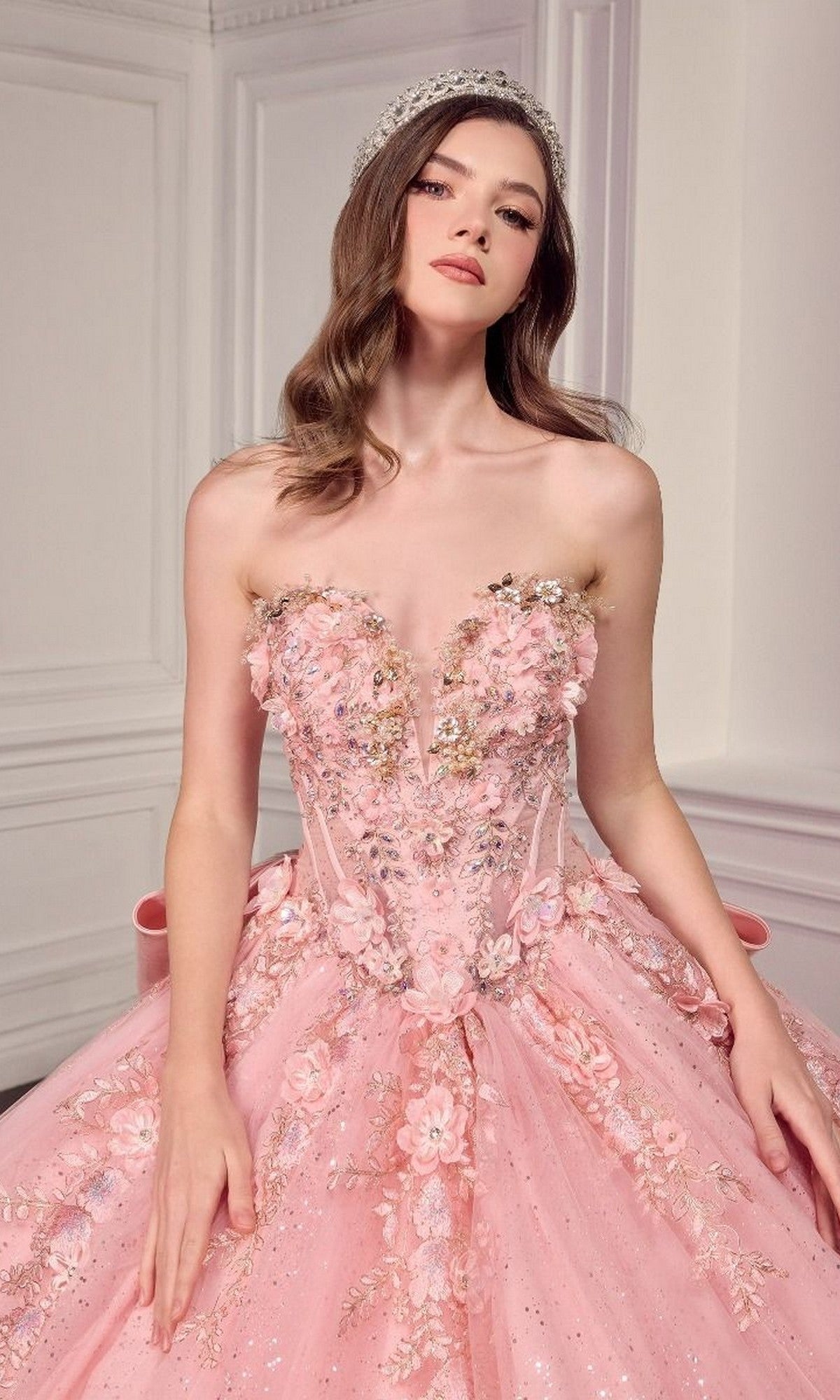 A woman in the Quinceanera Dress 15726 By Ladivine, featuring beaded 3D flowers and glitter tulle, stands in an elegant white-paneled room and completes her look with a sparkling tiara.